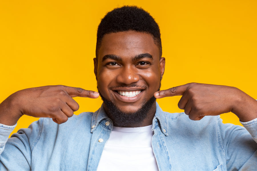 Dark-haired man against a yellow wall smiles and points to his teeth after professional teeth whitening in Martin, TN